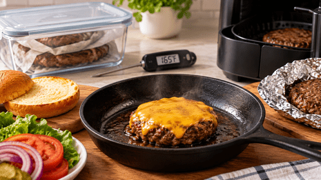 Juicy cheeseburger patty with melted cheddar cheese reheating in a cast-iron frying pan, with toasted burger buns, fresh lettuce, tomato, and a food thermometer nearby.